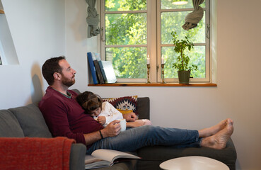 A father sitting on the sofa with his daughter sleeping on his lap on a relaxing afternoon at home.