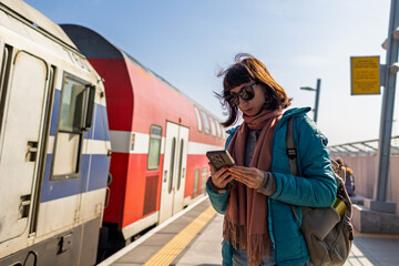 A young woman stands on the platform using her phone. A passenger checks the train schedule using an app on his phone. © zhukovvvlad