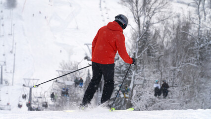 A skier in a red jacket stands on a picturesque ski slope, with a chairlift in the background and snow-covered trees framing the view. Snow dust flies up from under his skis.