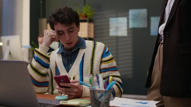 Bored young male employee distracted by smartphone scrolling content sitting at office desk with laptop while colleague scolding him for indiscipline