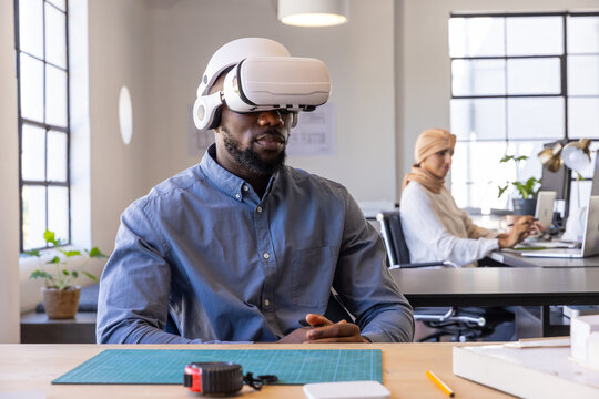 Diverse coworkers using vr headset and laptop at office desk with cutting mat, tape measure - Powered by Adobe