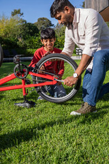Fototapeta premium Asian father and son kneeling on lawn near patio repairing red bicycle with wrench, gloves