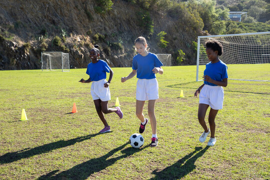 Diverse female teenage teammates dribbling soccer ball between cones on grassy field - Powered by Adobe