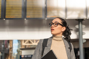Woman in glasses looks up at display board in busy train station during daylight hours