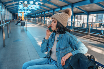 Woman sitting at a transport station talking on the phone while wearing winter clothing