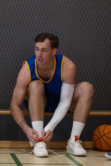 Naklejka premium Male basketball player sitting on wooden bench tying shoes in gym by basketball