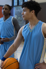 Diverse male teammates standing on gym court wearing jerseys and holding basketball