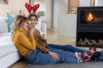 Naklejka premium Couple sitting on rug in living room near fireplace wearing antler headbands holding ceramic mugs