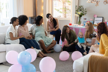 Diverse female friends celebrating baby shower in living room with balloons, baby, camera