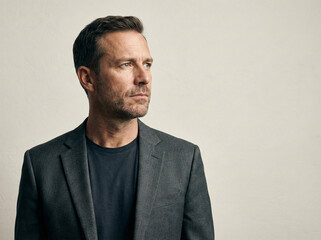 Obraz premium Middle aged man with stubble wearing grey suit jacket and dark t-shirt looking away thoughtfully