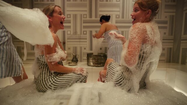 Women enjoy a traditional foam bath in a luxurious hammam, laughing and relaxing during a pampering spa treatment. Service staff assist them, creating a joyful and serene experience