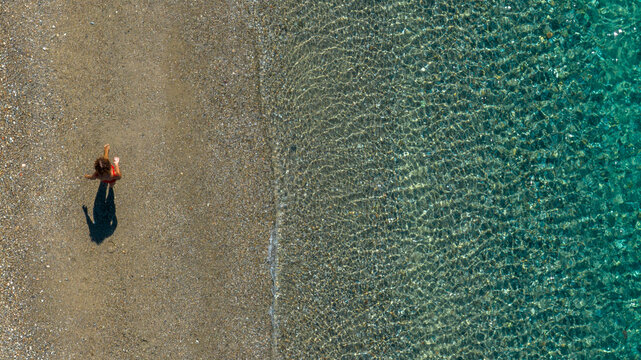 Top-down aerial shot of a lone person walking on a pebble beach. The crystal-clear shallow water creates a mesmerizing texture of ripples over the sandy seabed. Summer holiday concept. Zenithal view.