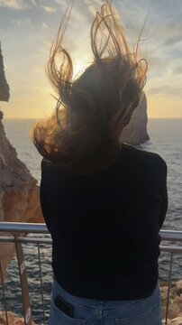 Cinematic slow motion video of a woman seen from behind as she stands at a panoramic viewpoint overlooking the sea. Strong wind moves her hair gracefully, creating dynamic and expressive motion 