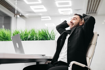 Male office worker in a black suit is celebrating success while sitting at a desk with a laptop, indoor plants visible in a modern office environment with bright lighting
