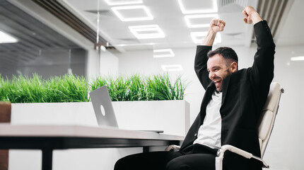 Male office worker in a black suit celebrates success with raised arms while sitting at a desk with a laptop and indoor plants in a modern office environment