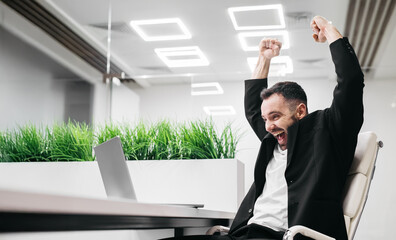 Male office worker in a black suit celebrating success with raised arms while sitting at a desk with a laptop and green plants in a modern workspace with bright lighting