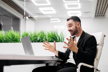 Male professional in black suit is engaged in a video call at a modern office with greenery in the background and bright overhead lighting during a business discussion