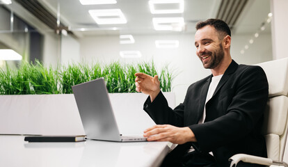 Male professional in black suit is sitting at a modern desk with a laptop, smiling and gesturing during a virtual meeting in a bright office environment with greenery