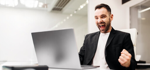 Male office worker in a black blazer is celebrating success while sitting at a desk with a laptop, bright modern workspace with glass walls and soft lighting visible in the background