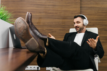 Businessman in a black suit with headphones is sitting at a desk with feet up, engaging in a virtual meeting on a laptop in a modern office environment with wooden accents