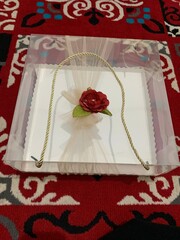 Clear box contains multiple small cakes  adorned with decorative element featuring red artificial flower and green leaves tied golden rope, suggesting wedding gift presentation.
