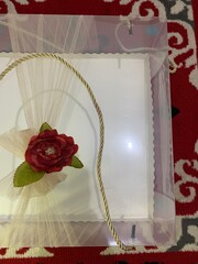 Clear box contains multiple small cakes  adorned with decorative element featuring red artificial flower and green leaves tied golden rope, suggesting wedding gift presentation.