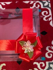 Clear box contains multiple small cakes  adorned with decorative element featuring red artificial flower and green leaves tied golden rope, suggesting wedding gift presentation.