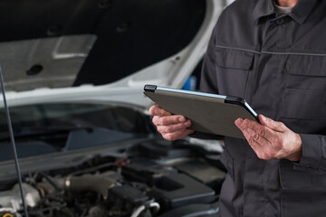 Midsection of man holding digital tablet inspecting car engine in auto repair shop, standing near open hood, wearing work uniform, focusing on diagnostic process