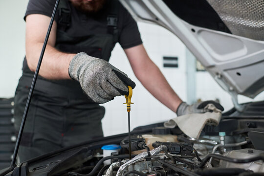 Midsection of man checking oil level in car engine bay, wearing work gloves and overalls, holding dipstick while performing maintenance under open hood in automotive workshop