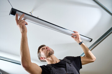 Man in workwear installing a linear LED light fixture on the ceiling indoors. Concept of electrical...