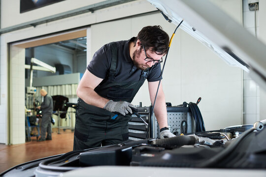Caucasian young adult man wearing glasses working on car engine in auto repair shop, using tool while inspecting under hood, focused on maintenance task in professional garage setting