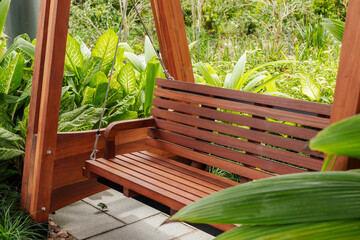 A wooden swing bench rests in a garden filled with various green plants. The setting is bright and sunny, showcasing natural surroundings
