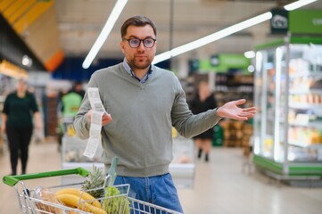 Shocked caucasian man with rounded eyes looking at extremely long bill. Amazed buyer doing grocery...