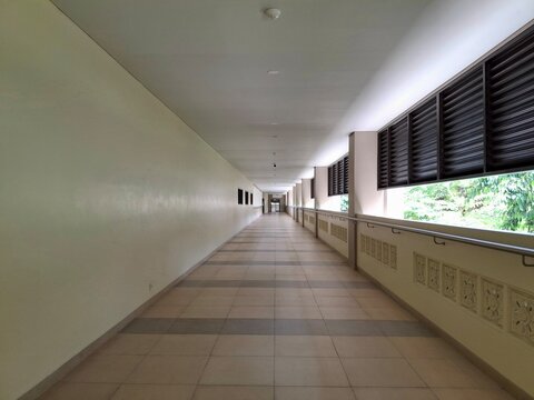 Long empty corridor perspective view in modern building. Indoor hallway architecture with tiled floor, ceiling lights, and windows with louvers leading to vanishing point.