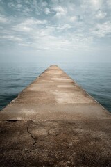 Fototapeta premium A weathered concrete pier stretches into a calm sea under a cloudy sky. Concept Weathered Pier, Calm Sea, Cloudy Sky, Moody Atmosphere, Nautical Photography