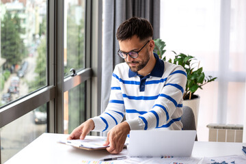 Side view of a modern professional man analyzing data on a laptop in a bright home workspace. Concept of digital transformation, hybrid work lifestyle, and successful small business management.