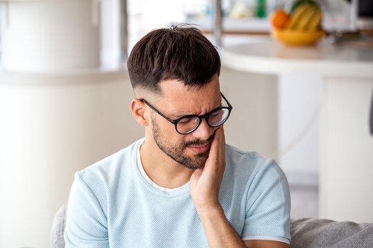 Young man in glasses suffering from severe toothache and jaw pain at home; concept for dental emergency, sensitive teeth, cavity, oral health, and need for professional dental care.