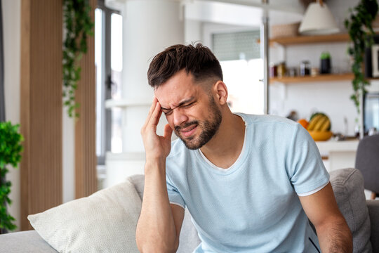 Young man holding his head in pain while suffering from a severe headache or migraine at home caused by stress or tension and high blood pressure while feeling fatigue and exhaustion