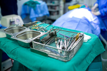 Metal tray with various sterile stainless steel surgical instruments and tools on a green medical table in a hospital operating room with a patient and surgical team in the blurred background