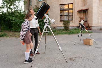 Man and a young girl stand next to a telescope outdoors, looking attentively as they prepare the optical instrument for celestial observation during an educational astronomy activity