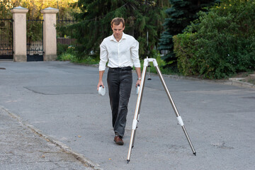 A man in a white shirt walks with telescope mount components in his hands, preparing for tripod assembly outdoors near stone structures and trees