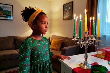 Obraz premium Black girl child standing beside kinara with seven lit candles during Kwanzaa celebration, gazing thoughtfully at candlelight in living room with festive decorations visible