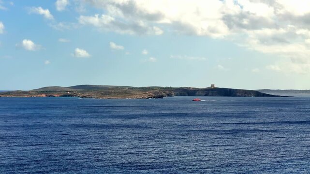 Red passenger ferry boat cruising through the turquoise North Comino Channel with the historic Saint Marys Tower on the rocky cliffs of Comino Island under a clear sunny sky in Malta during summer