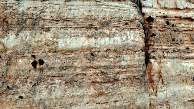 Deep eye level shot of a rugged limestone cliff face featuring dramatic horizontal sediment layers and vertical cracks under bright summer sun showing natural earth tones of beige tan and warm ochre
