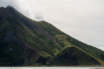 View of a green mountain slope covered with dense vegetation, partially shaded under cloudy sky, highlighting rugged ridges and natural landscape beauty. Sakhalin Island