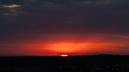 Fiery Red Sunset Over Distant City Horizon. A dramatic red and orange sunset glows beneath heavy clouds as the sun dips below the horizon, creating a powerful atmospheric landscape with intense colors