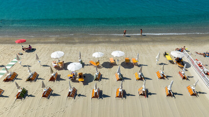 Aerial view of an organized beach. Rows of white sun umbrellas and orange lounge chairs line the sand, facing the calm, clear turquoise waters of the Mediterranean. Closeup from above of a beach club.