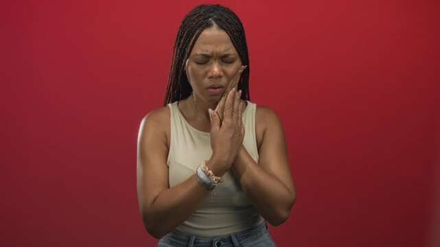 Young african american woman with hand on cheek for toothache in studio against red backdrop, jaw clenched and open mouth; pain discomfort.