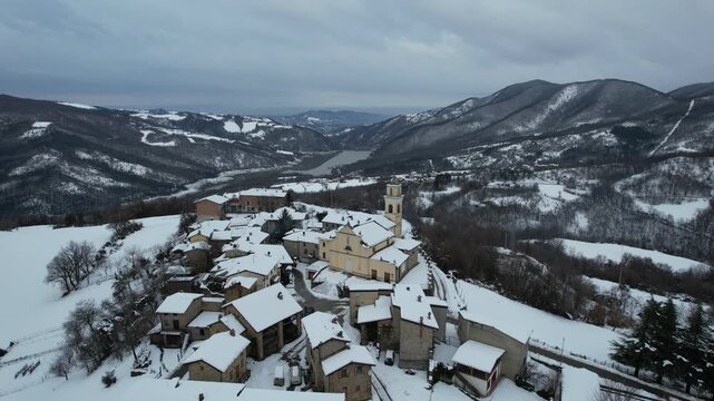 Drone establishing shot of snow covered Vezzolacca mountain village in the Piacenza Apennines, Emilia Romagna, Italy, showing snow covered mountain village, church bell tower and Mignano reservoir.
