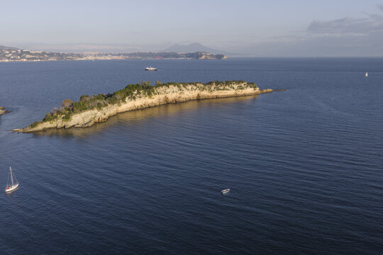 Aerial view of a rugged island with sparse vegetation amidst the vast, blue sea, a distant coastline and mountains adding depth, Capo Miseno, Bacoli, Italy.